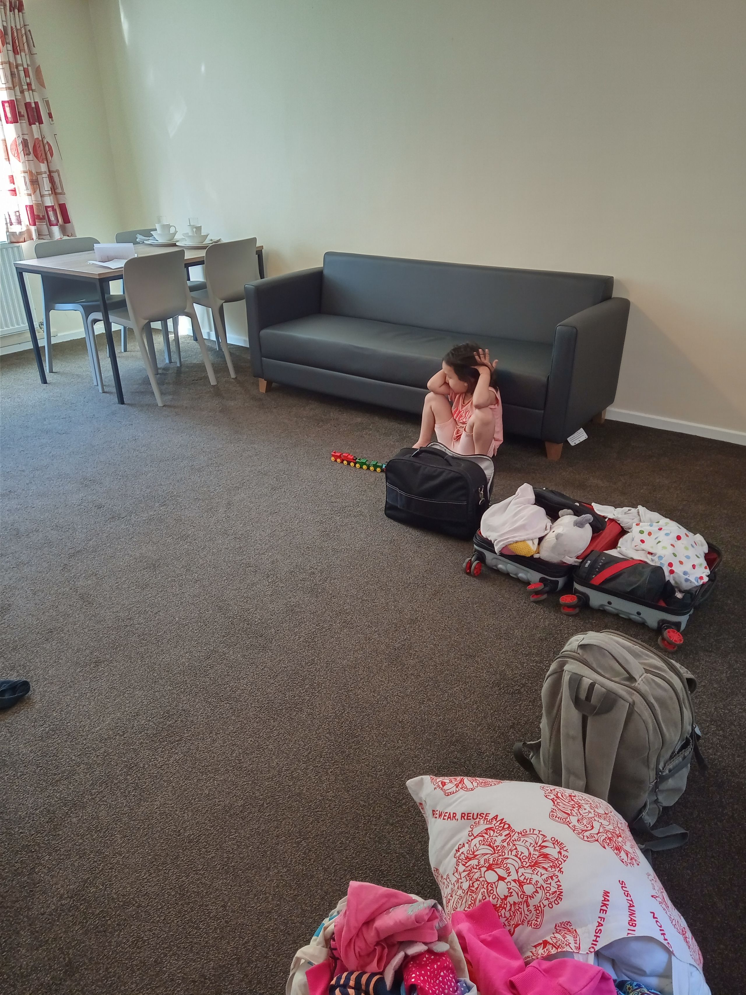 Portrait image of Ernest's UK accommodation. The image shows some personal items such as backpacks, cushions, open luggages and Ernest's young daughter sitting on the floor.